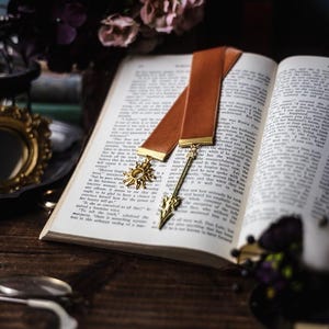 Puede incluir: Un marcapáginas de cuero marrón con un diseño de flecha y sol de oro. El marcapáginas está colocado en un libro abierto con páginas blancas.