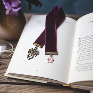 K&ouml;nnte beinhalten: Burgunderfarbenes Samt-Lesezeichen mit einem bronzefarbenen Drachenanh&auml;nger und einem rosa Bl&uuml;tenanh&auml;nger, das auf den offenen Seiten eines Buches liegt. Das Buch liegt auf einer Holzoberfl&auml;che, mit lila Blumen im Hintergrund.