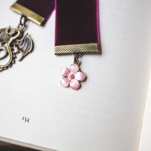 K&ouml;nnte beinhalten: Burgunderfarbenes Samt-Lesezeichen mit goldfarbenem Metallrand und rosa Bl&uuml;tenanh&auml;nger. Ein Drachenanh&auml;nger h&auml;ngt am anderen Ende. Das Lesezeichen liegt auf einer Buchseite mit der Nummer 134.