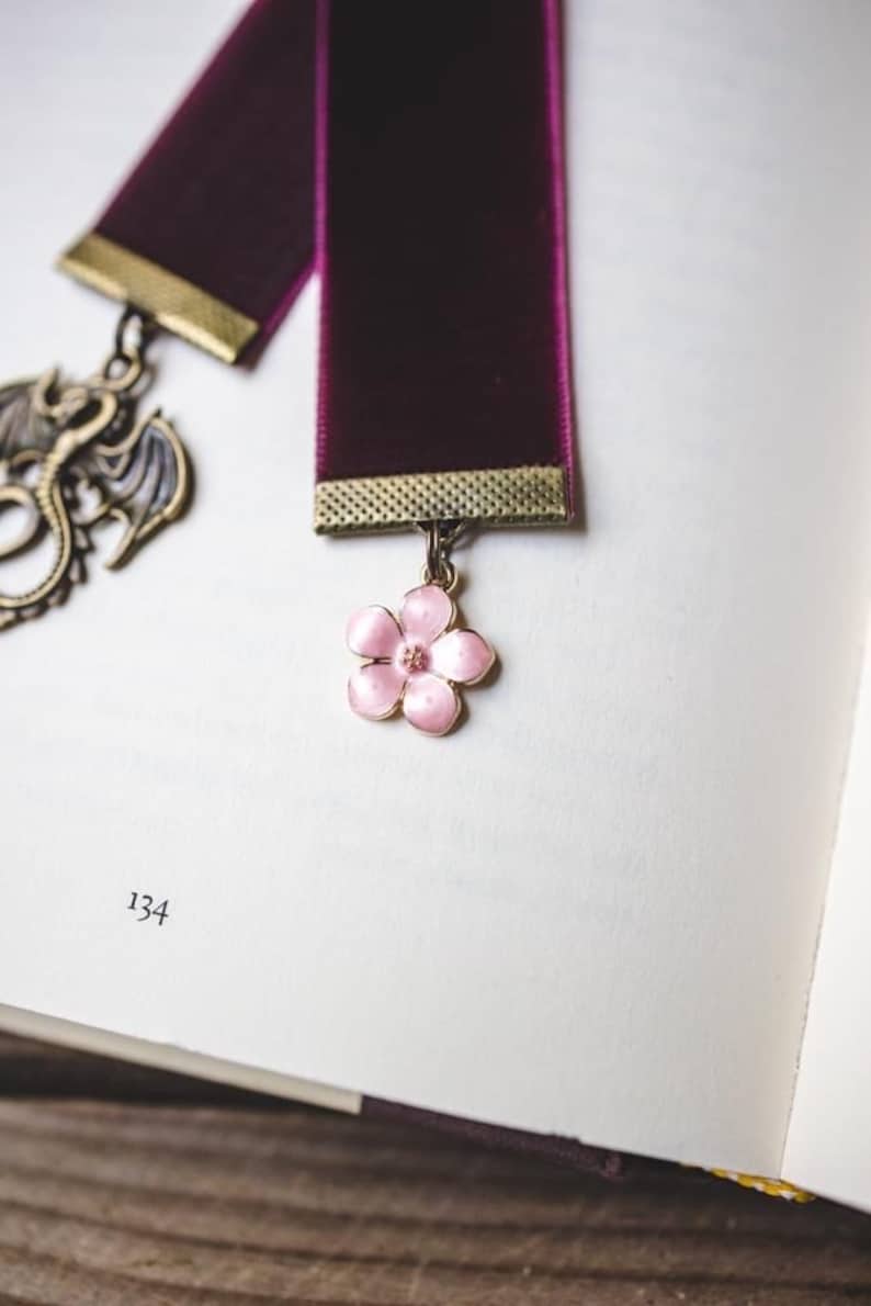 K&ouml;nnte beinhalten: Burgunderfarbenes Samt-Lesezeichen mit goldfarbenem Metallrand und rosa Bl&uuml;tenanh&auml;nger. Ein Drachenanh&auml;nger h&auml;ngt am anderen Ende. Das Lesezeichen liegt auf einer Buchseite mit der Nummer 134.