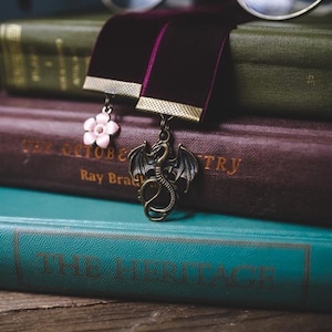 K&ouml;nnte beinhalten: Ein Stapel alter B&uuml;cher mit einer runden Brille darauf. Zwei Samt-Lesezeichen mit Anh&auml;ngern, eine rosa Blume und ein bronzener Drache, sind zwischen den Seiten platziert. Die B&uuml;cher sind in Gr&uuml;n, Burgunderrot und T&uuml;rkis gehalten.