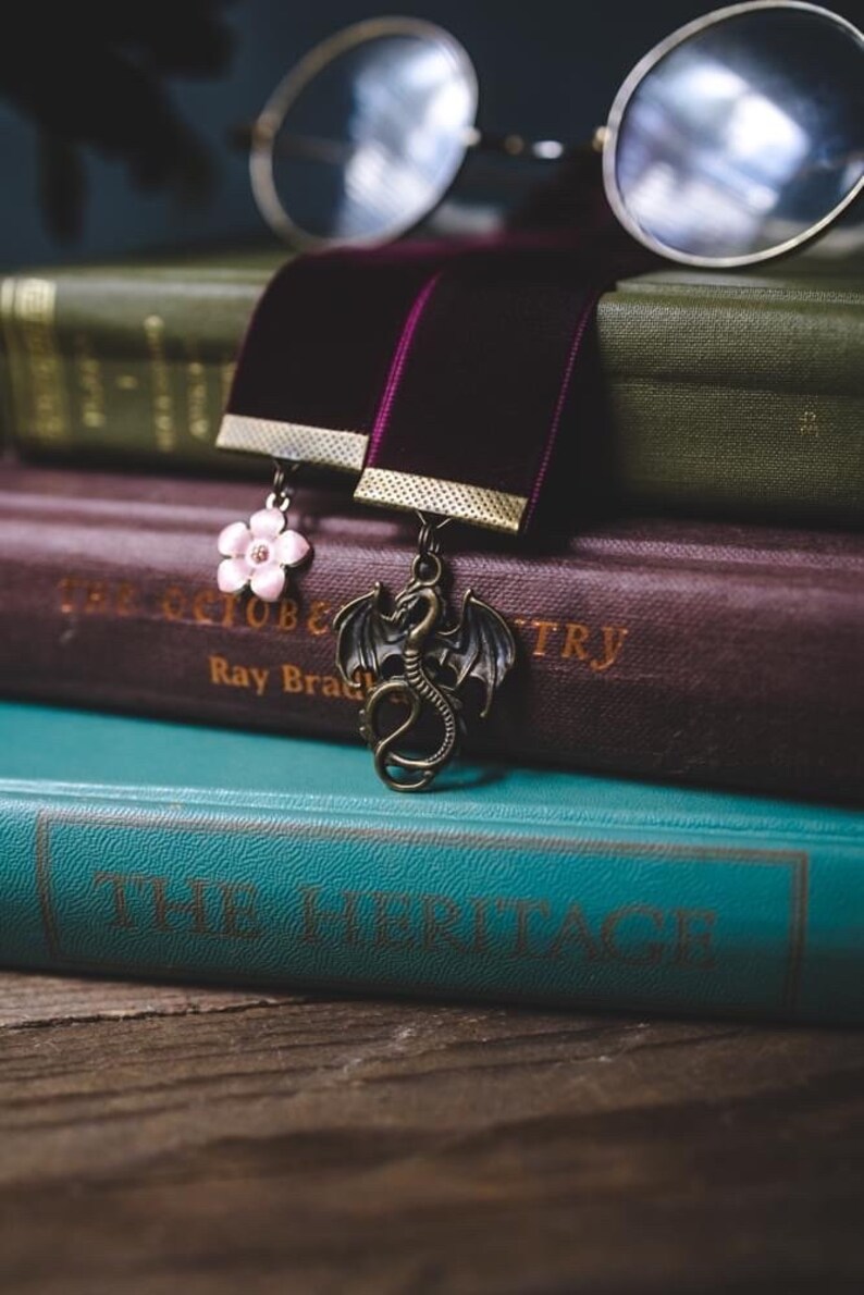 K&ouml;nnte beinhalten: Ein Stapel alter B&uuml;cher mit einer runden Brille darauf. Zwei Samt-Lesezeichen mit Anh&auml;ngern, eine rosa Blume und ein bronzener Drache, sind zwischen den Seiten platziert. Die B&uuml;cher sind in Gr&uuml;n, Burgunderrot und T&uuml;rkis gehalten.