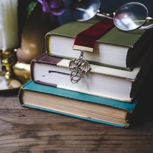 K&ouml;nnte beinhalten: Ein Stapel von drei B&uuml;chern mit einem roten Samt-Lesezeichen und einem drachenf&ouml;rmigen Buch-Anh&auml;nger. Das Lesezeichen h&auml;ngt am obersten Buch und der Anh&auml;nger h&auml;ngt am Lesezeichen. Ein Paar runde Brillen liegt auf den B&uuml;chern.