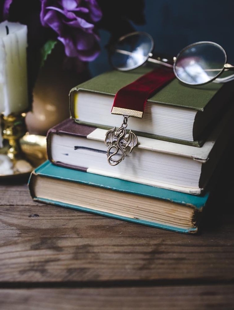 K&ouml;nnte beinhalten: Ein Stapel von drei B&uuml;chern mit einem roten Samt-Lesezeichen und einem drachenf&ouml;rmigen Buch-Anh&auml;nger. Das Lesezeichen h&auml;ngt am obersten Buch und der Anh&auml;nger h&auml;ngt am Lesezeichen. Ein Paar runde Brillen liegt auf den B&uuml;chern.