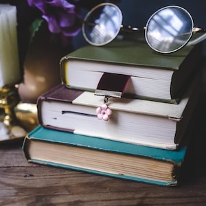 K&ouml;nnte beinhalten: Ein Stapel von drei alten B&uuml;chern mit runden Brillen obenauf. Ein Lesezeichen mit rosa Blumenanh&auml;nger h&auml;ngt am mittleren Buch. Die B&uuml;cher haben gr&uuml;ne, braune und t&uuml;rkisfarbene Einb&auml;nde. Eine Kerze und lila Blumen sind im Hintergrund.