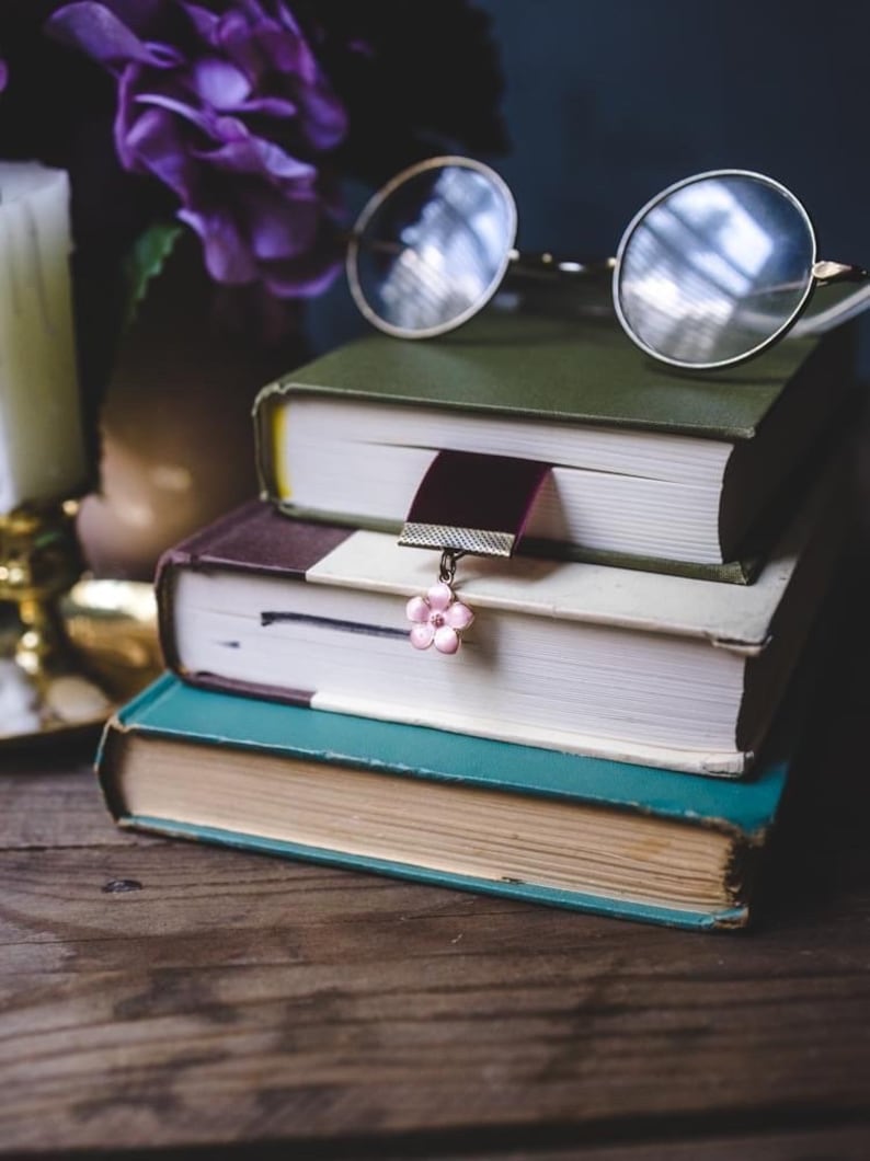 K&ouml;nnte beinhalten: Ein Stapel von drei alten B&uuml;chern mit runden Brillen obenauf. Ein Lesezeichen mit rosa Blumenanh&auml;nger h&auml;ngt am mittleren Buch. Die B&uuml;cher haben gr&uuml;ne, braune und t&uuml;rkisfarbene Einb&auml;nde. Eine Kerze und lila Blumen sind im Hintergrund.