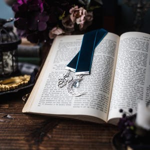 May include: A blue velvet bookmark with a silver dragon and crescent moon charm. The bookmark is placed in an open book with white pages.