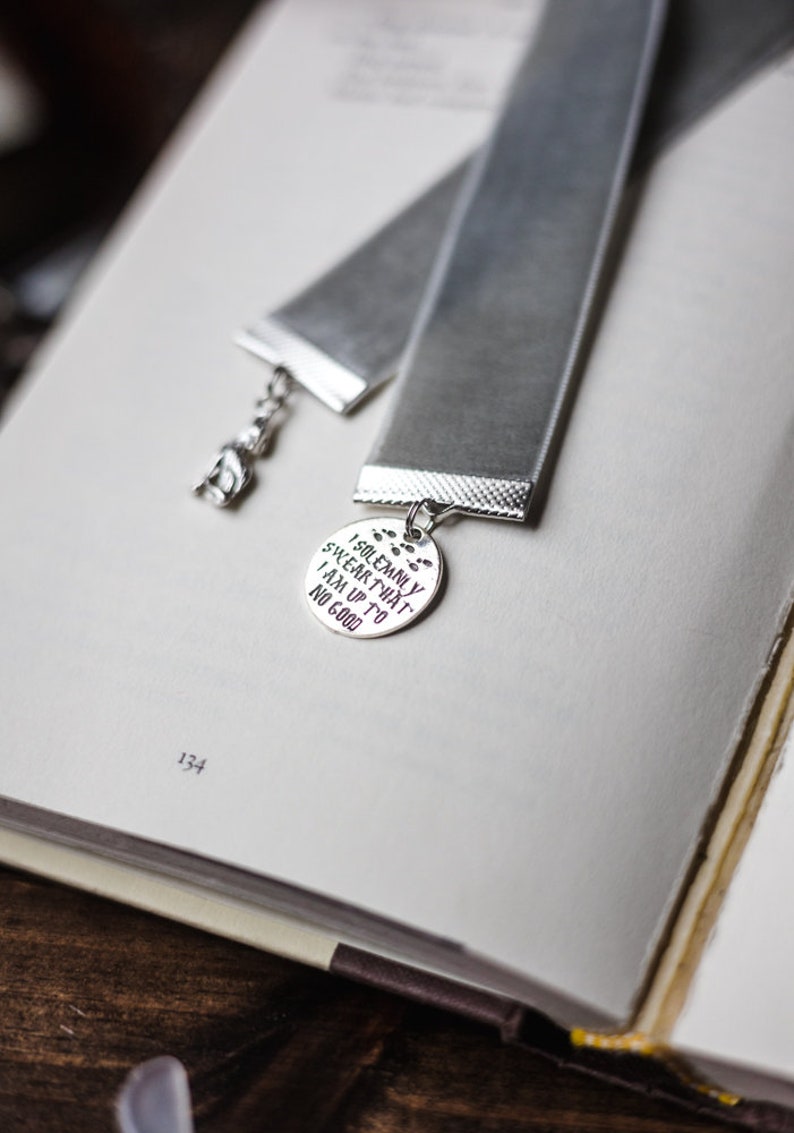 May include: A silver bookmark with a grey velvet ribbon. The bookmark features a round charm with the text "I solemnly swear that I am up to no good".