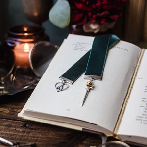 May include: A teal velvet bookmark with a silver heart charm and a silver sword charm with a gold crown. The bookmark is laying on an open book.