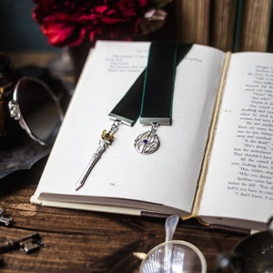 Puede incluir: Dos marcapáginas de terciopelo verde con detalles plateados y dorados. Un marcapáginas presenta una espada con una corona y el otro un círculo con una gema azul en el centro. Los marcapáginas están sobre un libro abierto con texto.