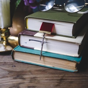 K&ouml;nnte beinhalten: Ein Stapel von vier B&uuml;chern mit einem roten Lesezeichen und einem silbernen Schwertlesezeichen. Die B&uuml;cher liegen auf einem Holztisch mit einer Kerze und Gl&auml;sern im Hintergrund.