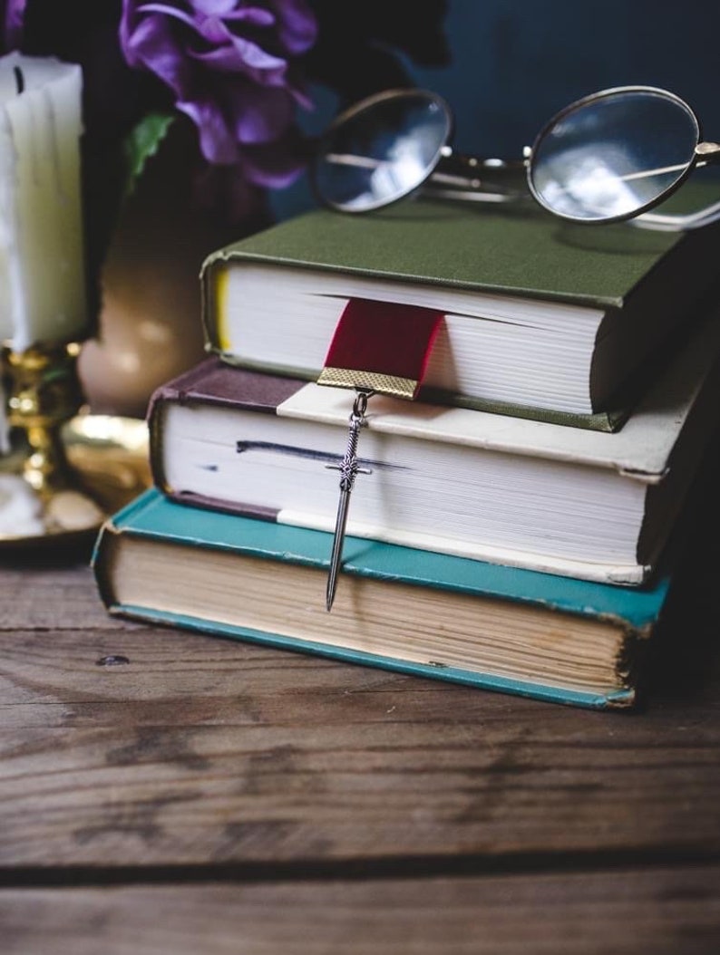 K&ouml;nnte beinhalten: Ein Stapel von vier B&uuml;chern mit einem roten Lesezeichen und einem silbernen Schwertlesezeichen. Die B&uuml;cher liegen auf einem Holztisch mit einer Kerze und Gl&auml;sern im Hintergrund.
