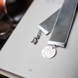 May include: A grey velvet bookmark with a silver charm attached. The charm features a small, detailed figure and a round disc with the text "I am envy, I am memory, And I am not, not real."