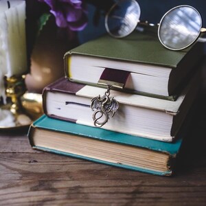 K&ouml;nnte beinhalten: Ein Stapel von drei Hardcover-B&uuml;chern mit gr&uuml;nen, braunen und t&uuml;rkisfarbenen Einb&auml;nden. Ein bronzener Drachenanh&auml;nger h&auml;ngt an einem weinroten Lesezeichen. Eine runde Brille liegt auf den B&uuml;chern.