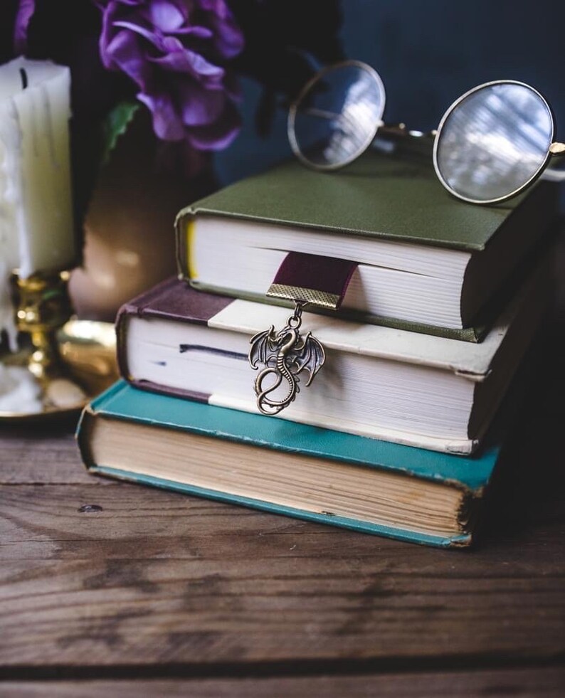 K&ouml;nnte beinhalten: Ein Stapel von drei Hardcover-B&uuml;chern mit gr&uuml;nen, braunen und t&uuml;rkisfarbenen Einb&auml;nden. Ein bronzener Drachenanh&auml;nger h&auml;ngt an einem weinroten Lesezeichen. Eine runde Brille liegt auf den B&uuml;chern.