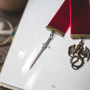 K&ouml;nnte beinhalten: Ein rotes Band Lesezeichen mit einem silbernen Schwert und einem goldenen Drachen-Anh&auml;nger. Das Lesezeichen liegt auf einem wei&szlig;en Buch.