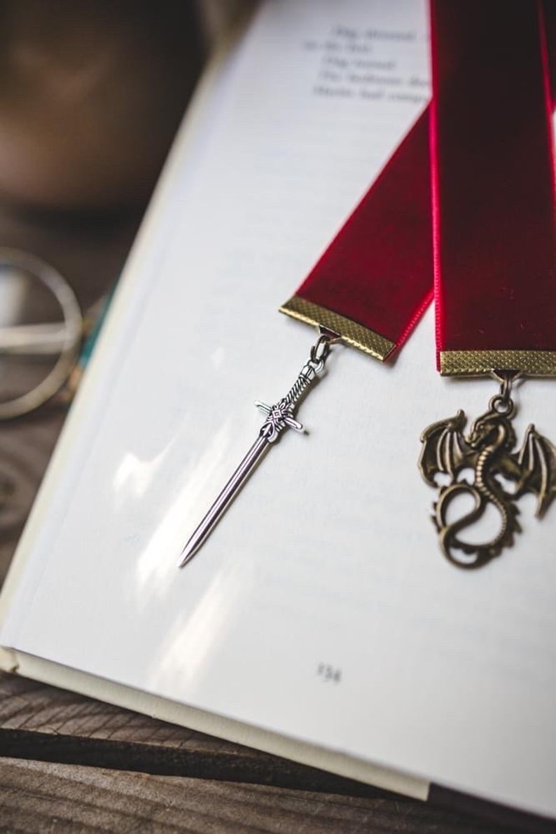 K&ouml;nnte beinhalten: Ein rotes Band Lesezeichen mit einem silbernen Schwert und einem goldenen Drachen-Anh&auml;nger. Das Lesezeichen liegt auf einem wei&szlig;en Buch.