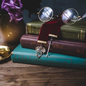 K&ouml;nnte beinhalten: Ein Stapel B&uuml;cher mit einem roten Samt-Lesezeichen und einem Drachen- und Schwertanh&auml;nger. Das Lesezeichen ist in das oberste Buch gesteckt, das gr&uuml;n ist und den Titel "Plato I" auf dem R&uuml;cken tr&auml;gt. Das zweite Buch ist braun und tr&auml;gt den Titel "The October Country" auf dem R&uuml;cken. Das unterste Buch ist t&uuml;rkisfarben und tr&auml;gt den Titel "The Heritage" auf dem R&uuml;cken.