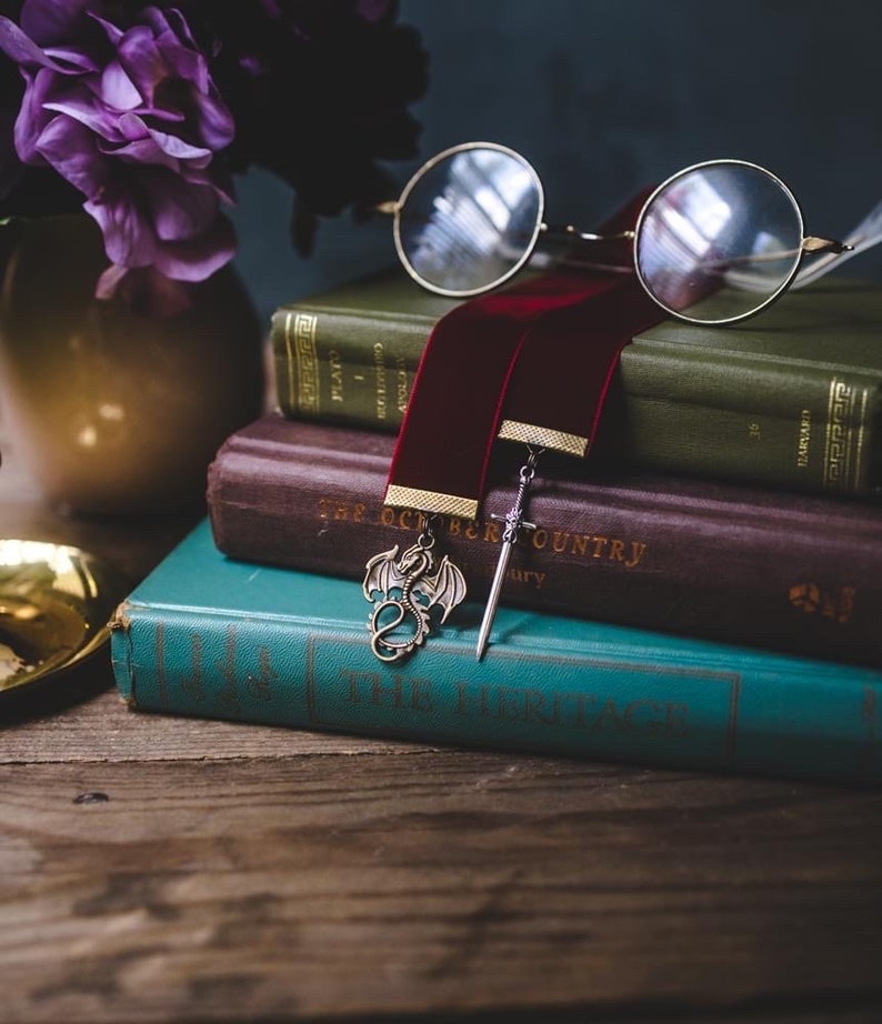 K&ouml;nnte beinhalten: Ein Stapel B&uuml;cher mit einem roten Samt-Lesezeichen und einem Drachen- und Schwertanh&auml;nger. Das Lesezeichen ist in das oberste Buch gesteckt, das gr&uuml;n ist und den Titel "Plato I" auf dem R&uuml;cken tr&auml;gt. Das zweite Buch ist braun und tr&auml;gt den Titel "The October Country" auf dem R&uuml;cken. Das unterste Buch ist t&uuml;rkisfarben und tr&auml;gt den Titel "The Heritage" auf dem R&uuml;cken.
