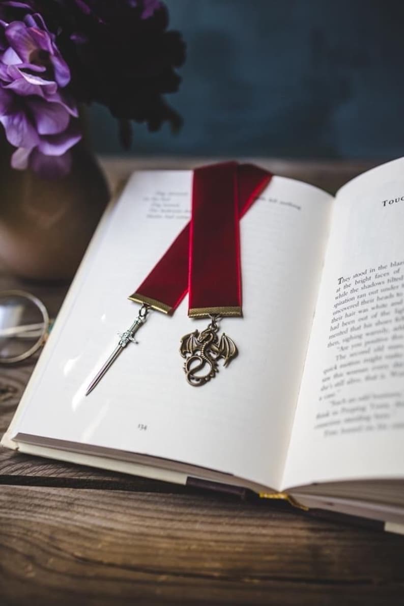 K&ouml;nnte beinhalten: Ein rotes Samt Lesezeichen mit einem silbernen Schwert und einem goldenen Drachen-Anh&auml;nger. Das Lesezeichen liegt auf einem ge&ouml;ffneten Buch mit wei&szlig;en Seiten.