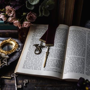 May include: An open book with a burgundy ribbon bookmark featuring a dragon charm and a sword charm. A bouquet of dark purple flowers and vintage books are in the background. Antique keys and a magnifying glass are scattered around the book.