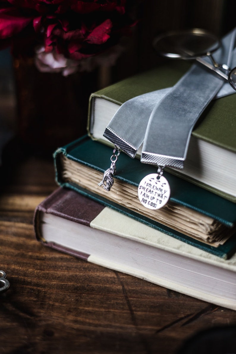 May include: A stack of three books with a grey velvet ribbon bookmark. The bookmark has two charms, one is a small figure and the other is a round charm with the text "I solemnly swear that I am up to no good".