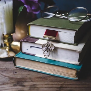 K&ouml;nnte beinhalten: Ein Stapel von drei B&uuml;chern mit einem roten Lesezeichen und einem drachenf&ouml;rmigen Lesezeichen-Anh&auml;nger. Die B&uuml;cher liegen auf einer Holzoberfl&auml;che mit einer Kerze und einer Brille im Hintergrund.