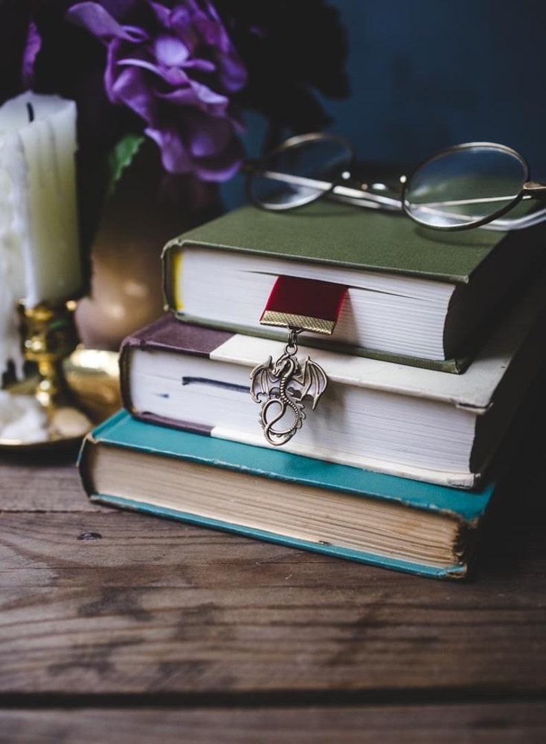 K&ouml;nnte beinhalten: Ein Stapel von drei B&uuml;chern mit einem roten Lesezeichen und einem drachenf&ouml;rmigen Lesezeichen-Anh&auml;nger. Die B&uuml;cher liegen auf einer Holzoberfl&auml;che mit einer Kerze und einer Brille im Hintergrund.