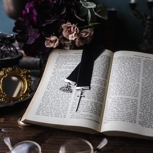May include: An open book with a black velvet bookmark featuring a silver cross and a decorative charm. A pair of round eyeglasses rests on the wooden surface. A bouquet of dark purple and pink flowers is in the background.