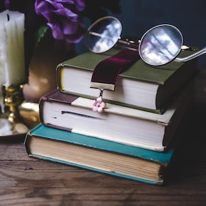 K&ouml;nnte beinhalten: Ein Stapel von drei alten B&uuml;chern mit unterschiedlich farbigen Einb&auml;nden. Ein burgunderfarbenes Samtlesezeichen mit einem rosa Blumenanh&auml;nger liegt zwischen den Seiten. Eine runde Brille liegt auf dem obersten Buch. Eine Kerze und lila Blumen sind im Hintergrund.