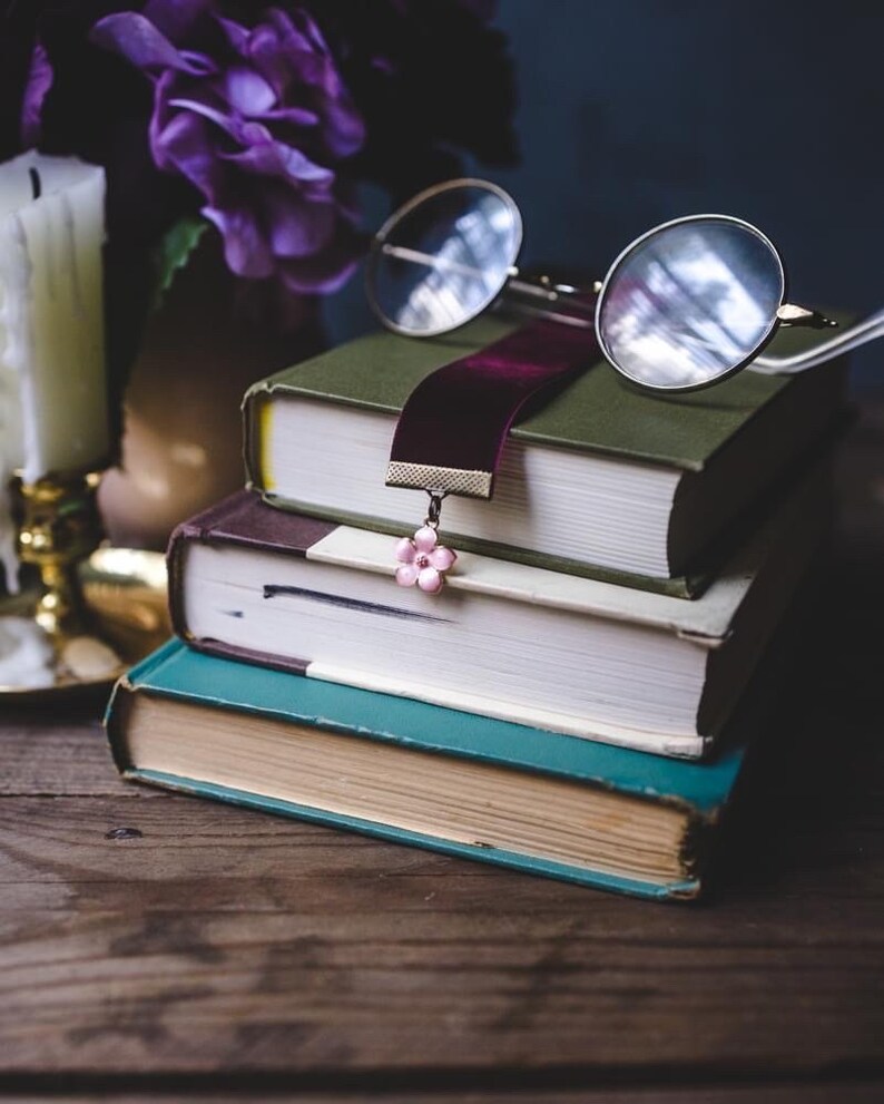 K&ouml;nnte beinhalten: Ein Stapel von drei alten B&uuml;chern mit unterschiedlich farbigen Einb&auml;nden. Ein burgunderfarbenes Samtlesezeichen mit einem rosa Blumenanh&auml;nger liegt zwischen den Seiten. Eine runde Brille liegt auf dem obersten Buch. Eine Kerze und lila Blumen sind im Hintergrund.