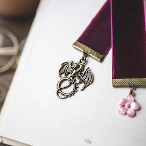 K&ouml;nnte beinhalten: Zwei burgunderfarbene Samtbuchzeichen mit dekorativen Anh&auml;ngern. Eines hat einen bronzenen Drachenanh&auml;nger, das andere eine rosa Blume. Die Lesezeichen liegen auf den offenen Seiten eines Buches, wobei die Seitenzahl 134 sichtbar ist.