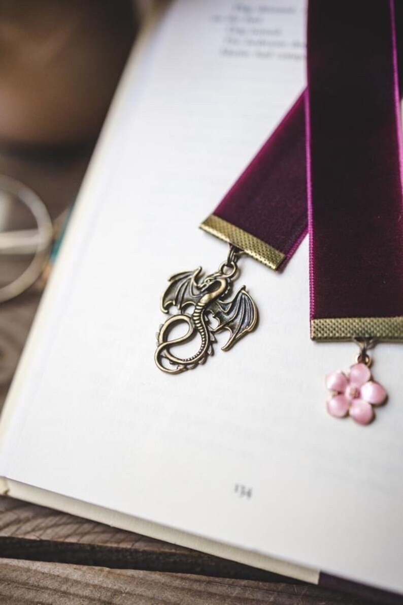 K&ouml;nnte beinhalten: Zwei burgunderfarbene Samtbuchzeichen mit dekorativen Anh&auml;ngern. Eines hat einen bronzenen Drachenanh&auml;nger, das andere eine rosa Blume. Die Lesezeichen liegen auf den offenen Seiten eines Buches, wobei die Seitenzahl 134 sichtbar ist.