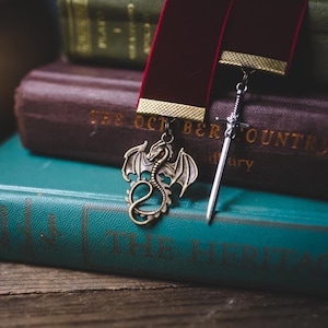 K&ouml;nnte beinhalten: Zwei Lesezeichen, eines mit einem Drachenmotiv und das andere mit einem Schwertmotiv, liegen auf einem Stapel B&uuml;cher. Das oberste Buch ist blaugr&uuml;n und hat den Titel "The Hertitage" auf dem Einband. Das darunterliegende Buch ist braun und hat den Titel "The October Country" auf dem Einband.