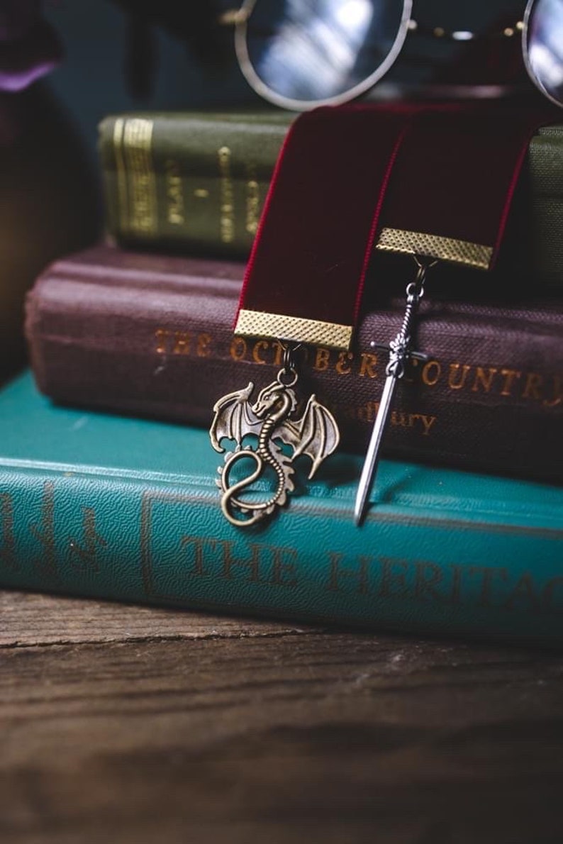 K&ouml;nnte beinhalten: Zwei Lesezeichen, eines mit einem Drachenmotiv und das andere mit einem Schwertmotiv, liegen auf einem Stapel B&uuml;cher. Das oberste Buch ist blaugr&uuml;n und hat den Titel "The Hertitage" auf dem Einband. Das darunterliegende Buch ist braun und hat den Titel "The October Country" auf dem Einband.