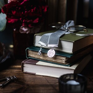 May include: A stack of three books with a grey velvet ribbon bookmark. The bookmark has two silver charms, one with a bird and the other with the text "Sometimes everything can add up to no good".