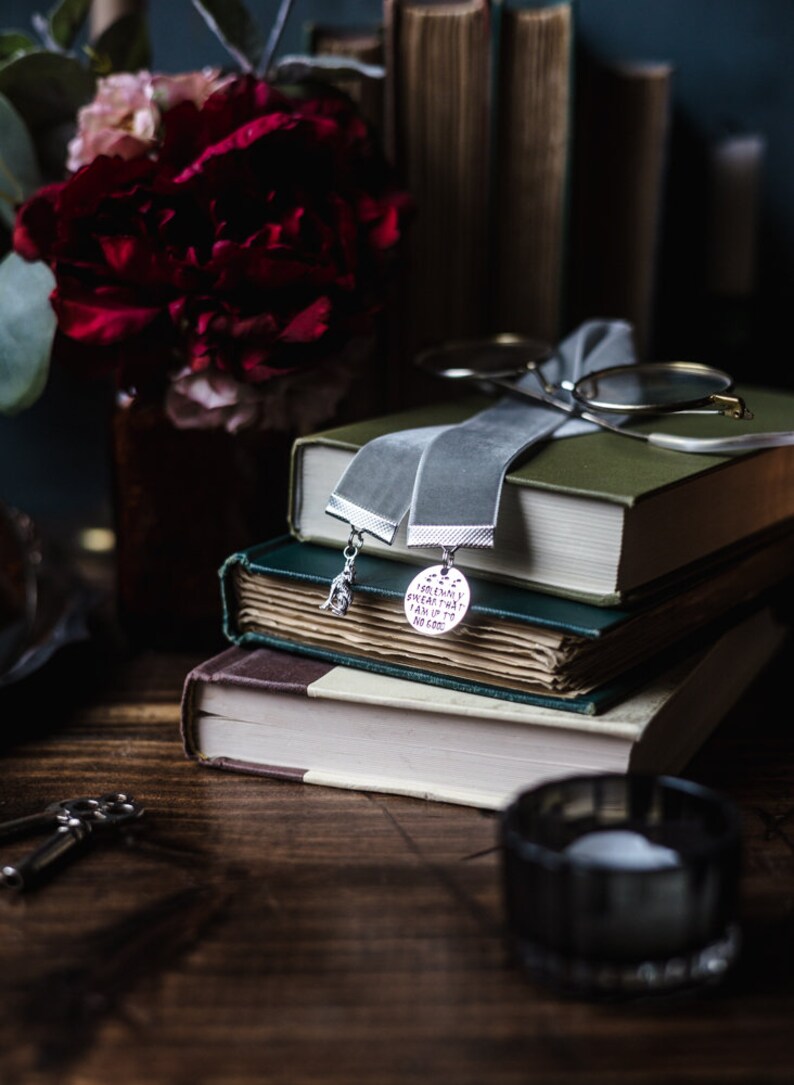 May include: A stack of three books with a grey velvet ribbon bookmark. The bookmark has two silver charms, one with a bird and the other with the text "Sometimes everything can add up to no good".