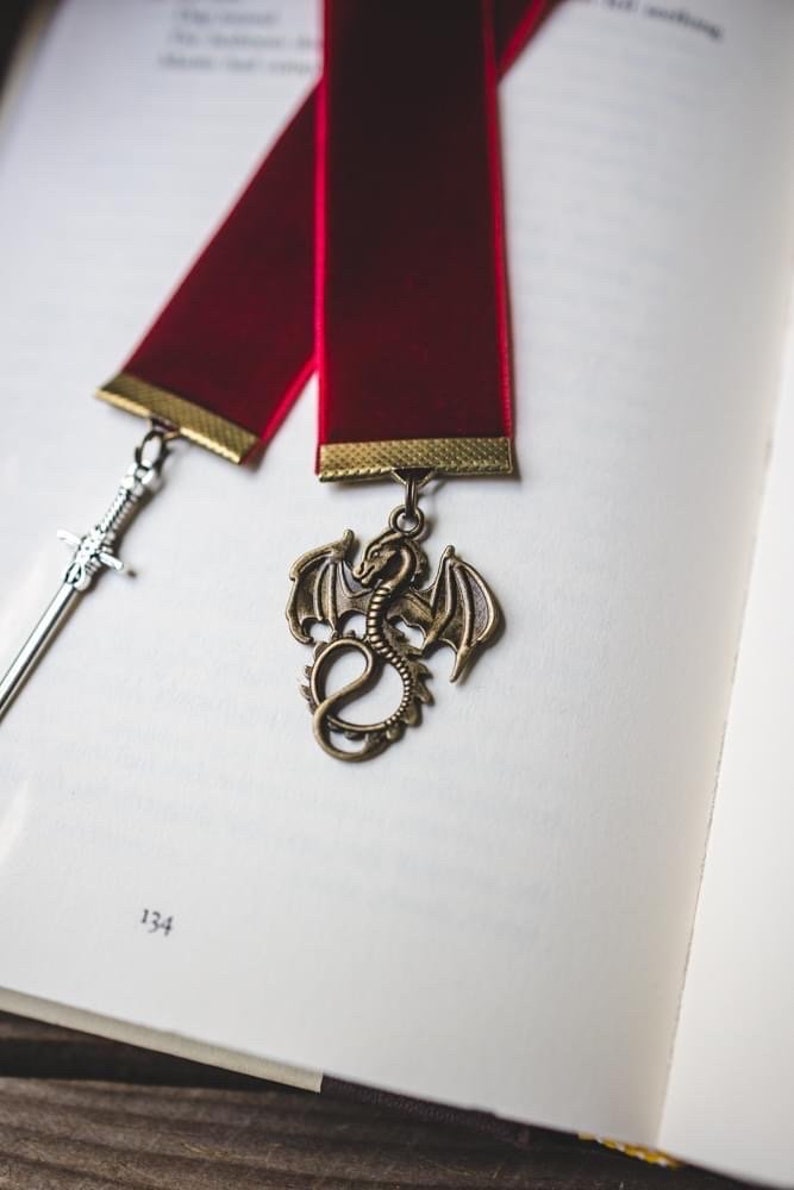 K&ouml;nnte beinhalten: Ein rotes Samt Lesezeichen mit goldener Einfassung und einem Drachen-Anh&auml;nger. Das Lesezeichen liegt auf einer wei&szlig;en Seite mit der Nummer 134 darauf.