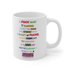 May include: White ceramic mug with a black handle. The mug features colorful text that reads "A FUCK MUST BE EARNED. I CAN'T GO DOWN THE STREET WITH A BUCKET OF FUCKS, GIVING THEM OUT TO EVERYONE. IF I FEEL THAT YOU'VE EARNED IT, I WILL GIVE A FUCK."
