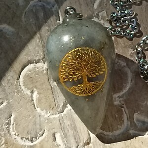 Op de afbeelding: Een grijze stenen slinger met een gouden levensboomontwerp. De slinger is bevestigd aan een zilveren ketting met een kleine grijze stenen kraal.