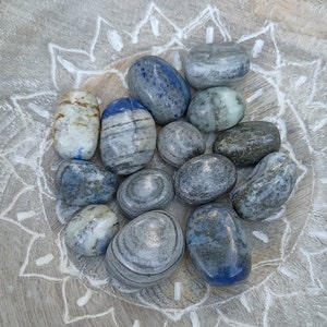 Puede incluir: Una colección de piedras pulidas en tonos grises y azules. Las piedras tienen un patrón estriado y están dispuestas en círculo sobre una superficie de madera.