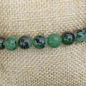 Puede incluir: Un brazalete de piedras preciosas verdes y negras con cuentas redondas. El brazalete está sobre un fondo de tela marrón.