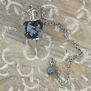 Puede incluir: Un péndulo plateado con una cadena y una piedra redonda moteada en blanco y negro. El péndulo tiene una punta y una pequeña piedra redonda al final de la cadena. El fondo es una superficie de madera clara tallada.