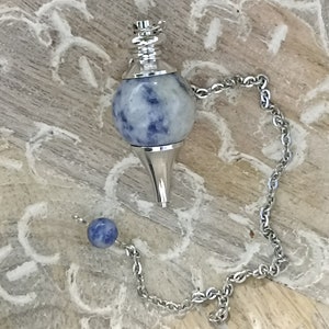 Può includere: Un pendolo di cristallo blu e argento con una catena. Il pendolo è rotondo con una punta appuntita e ha una sfera di cristallo blu e bianca.