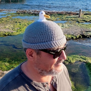 May include: A gray crocheted beanie hat with a white seagull figure on top. The seagull has a yellow beak. The hat is worn by a person wearing sunglasses. The background shows a rocky shoreline and ocean.
