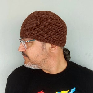 Peut inclure: Un bonnet en crochet marron. Le chapeau est fait d'un fil texturé et tissé serré. Le bonnet est de couleur marron chaud et uni et a une forme arrondie. Le chapeau est porté par une personne portant des lunettes et un t-shirt noir.