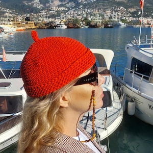 May include: A bright red knitted beanie hat with a small loop on top. The hat is worn with sunglasses that have the word "Vogue" printed on the side. The background shows boats on the water and a city in the distance.