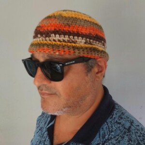 May include: A crocheted beanie cap featuring horizontal stripes in shades of orange, brown, yellow, and beige. The hat has a rounded crown and a short, slightly turned-up brim, constructed with a visible yarn stitch pattern throughout the fabric.