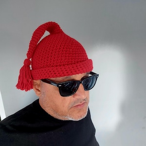 May include: A bright red crocheted beanie with a folded brim and a long, pointed top section that hangs down to the side, finished with a decorative yarn tassel at the end. The hat features a dense, textured stitch pattern throughout the design.