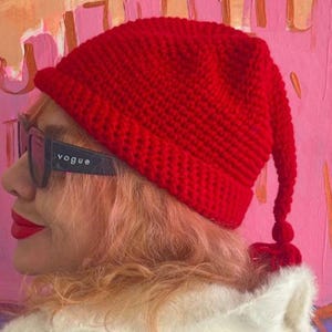 May include: A bright red crocheted beanie with a folded brim and a long, pointed tail that ends in a small tassel. The hat features a textured, chunky knit pattern throughout. The beanie is worn with dark-framed sunglasses with the word Vogue on them.